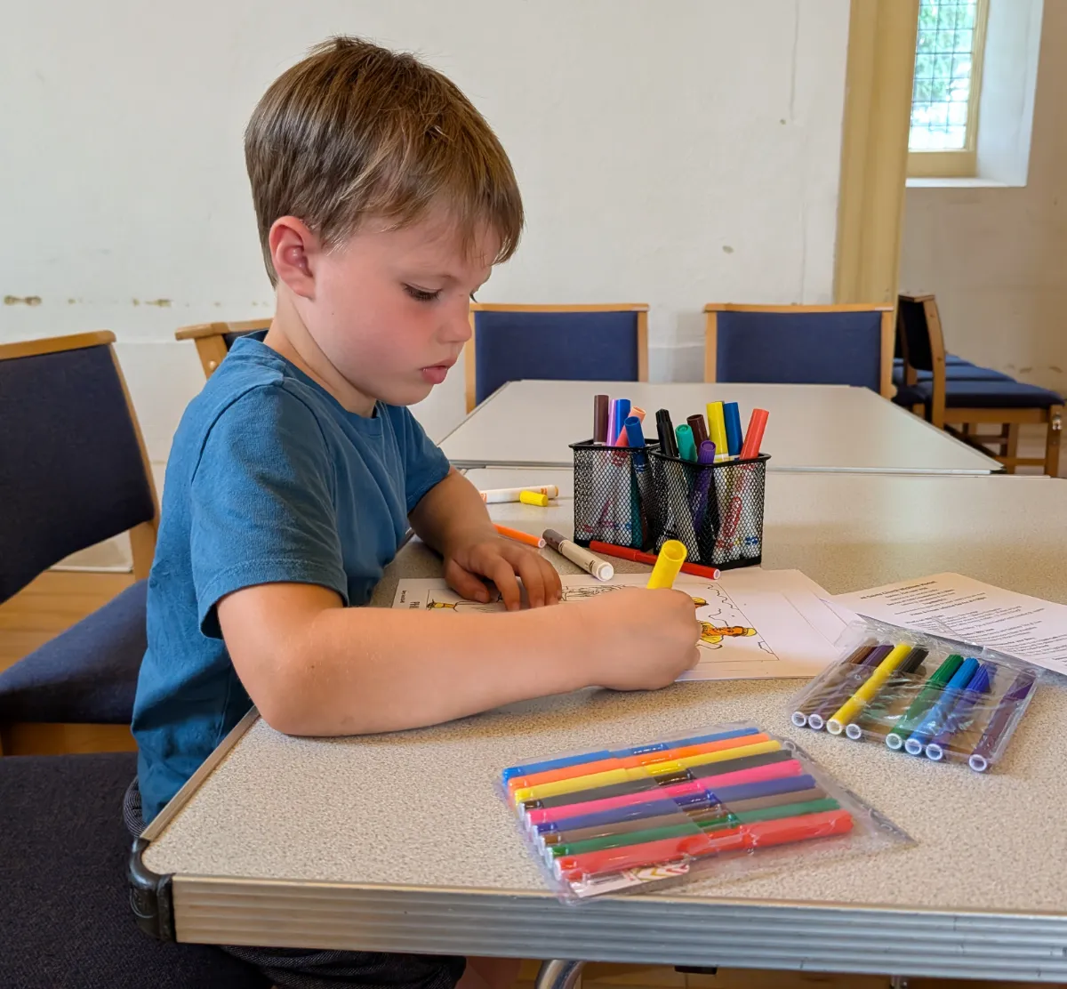 The brother at a long table, head down, drawing with felt-tip pens